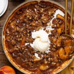 Peach crisp in a skillet with serving spoons, a scoop of ice cream, and peaches.