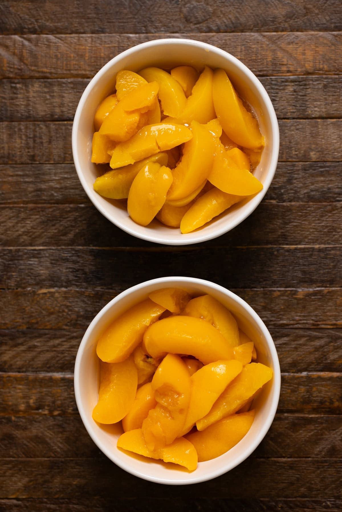 Sliced peaches in two white bowls on a brown wood table.