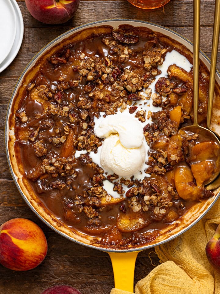 Peach crisp in a skillet with serving spoons, a scoop of ice cream, and peaches.