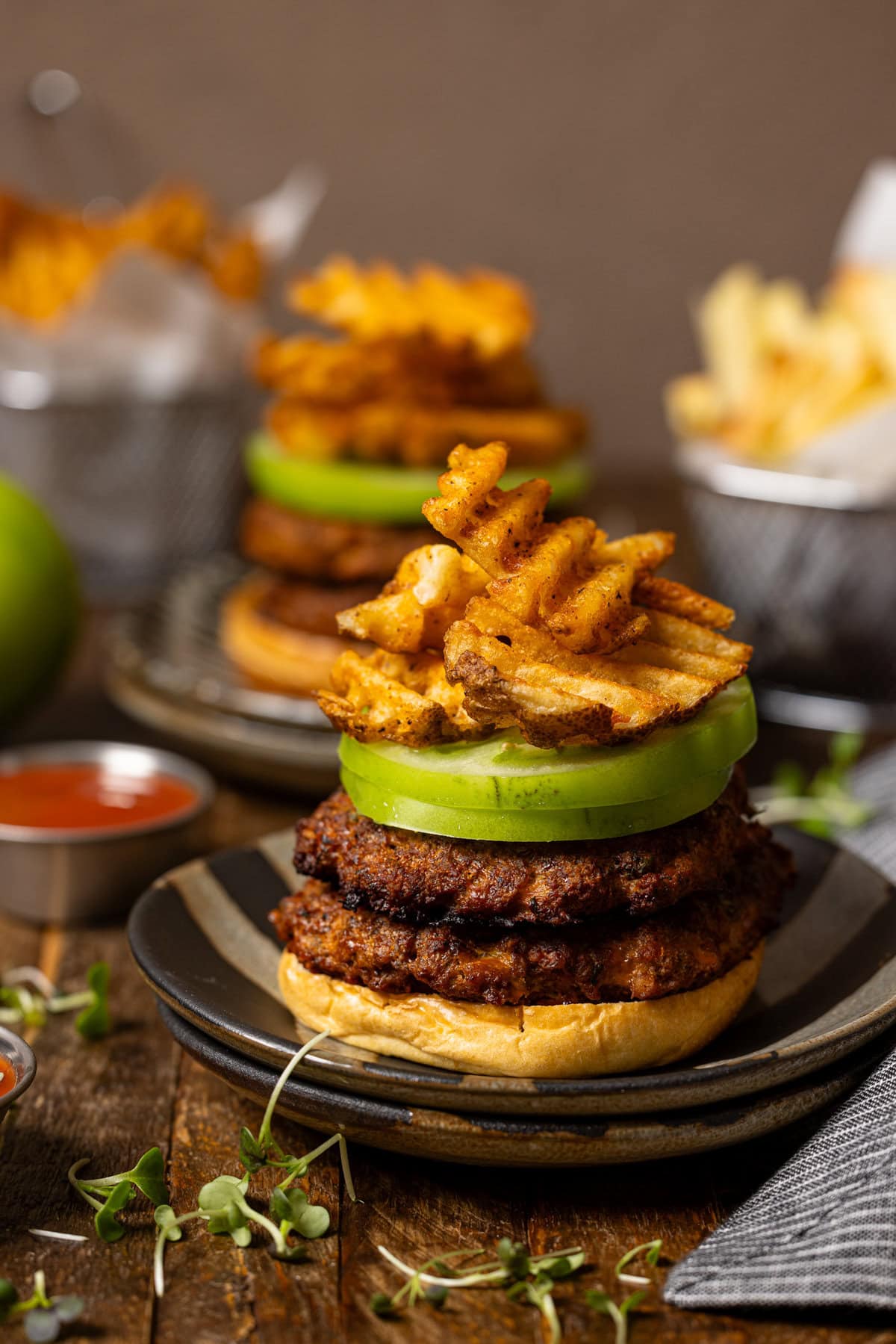 Burgers with toppings stacked and sauces and fries on the side. 