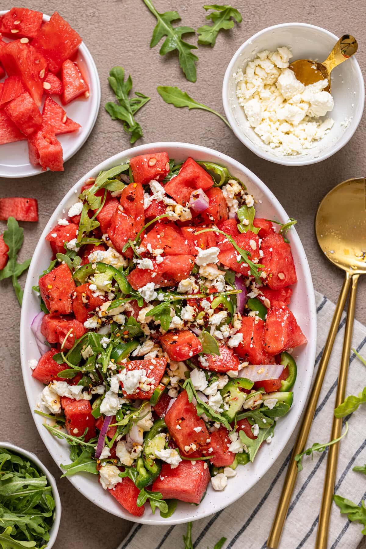 Salad in a white serving dish with gold spoons, chopped watermelon and feta cheese.