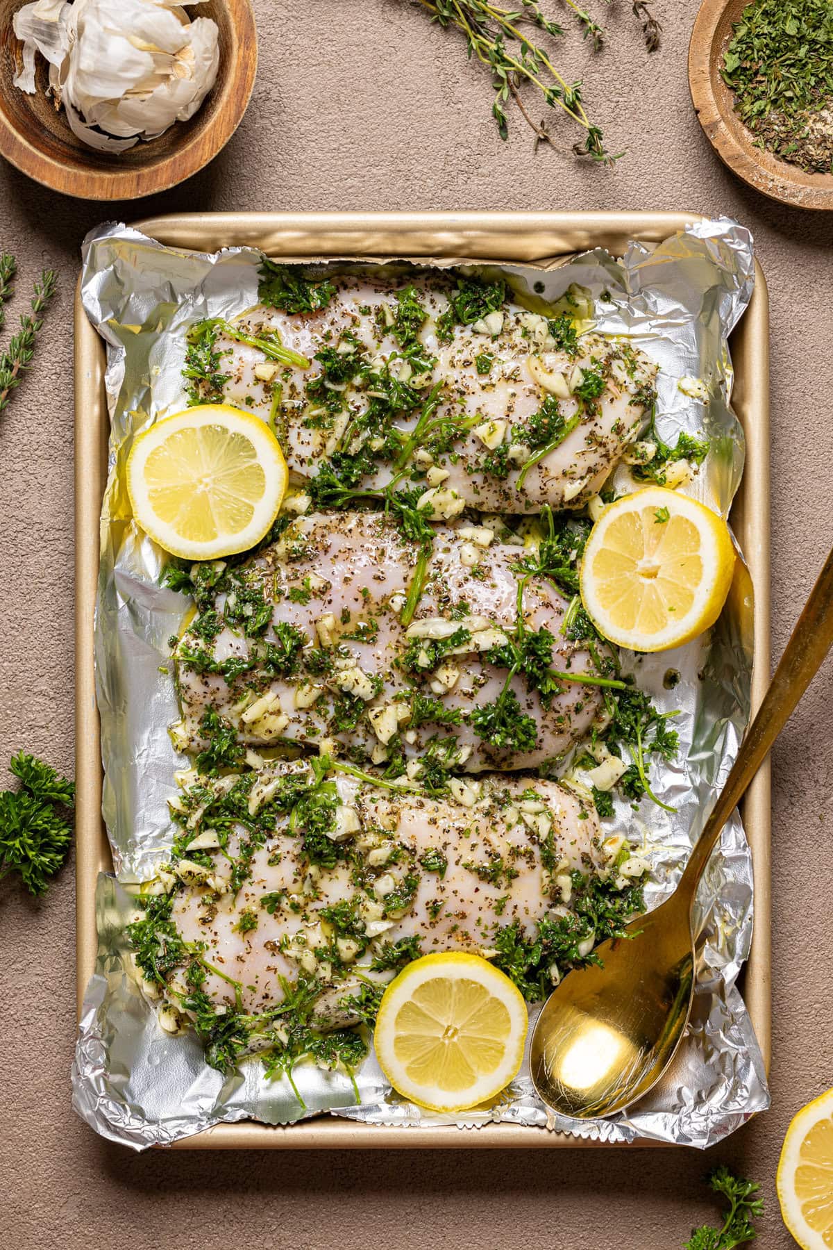 Marinated chicken on a baking sheet with foil paper and a gold spoon.