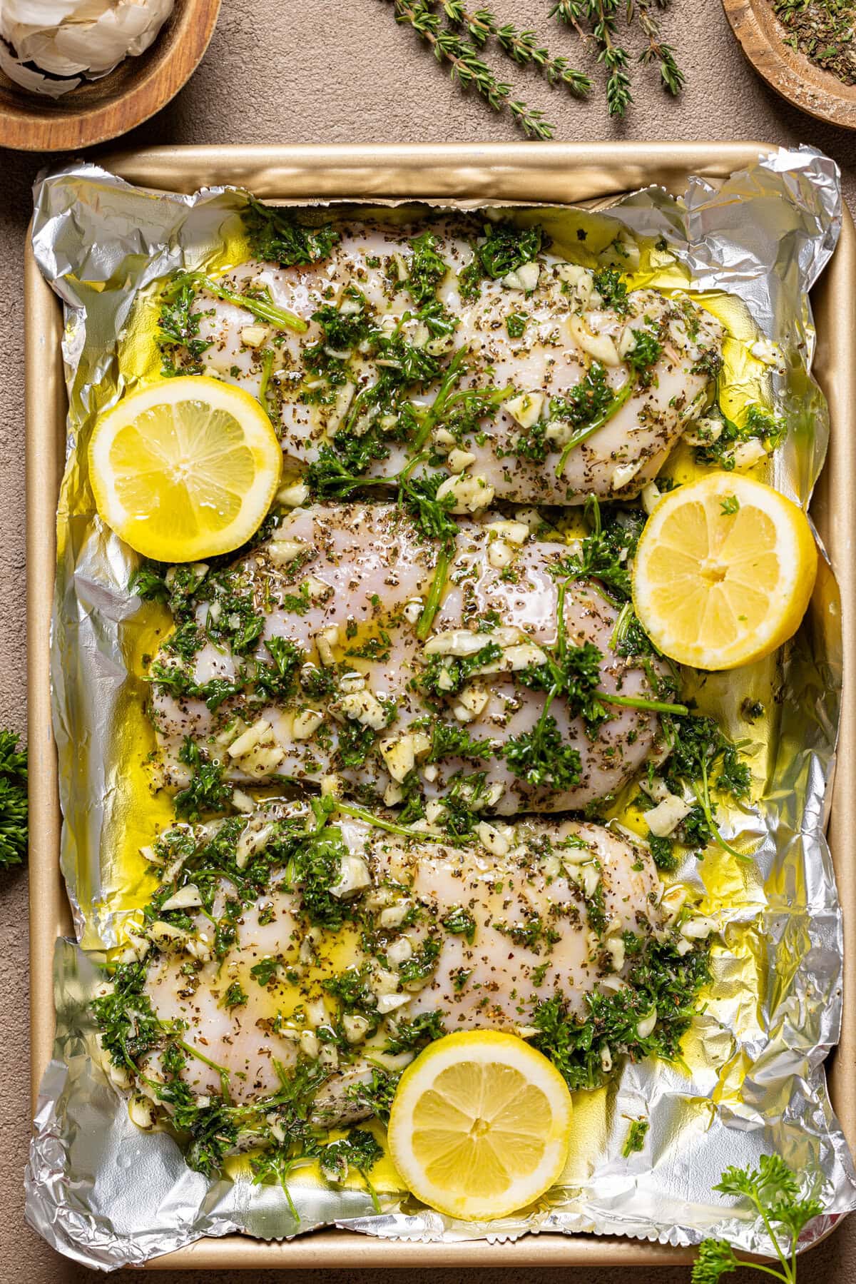 Marinated chicken on a baking sheet with foil, herbs, and lemons.