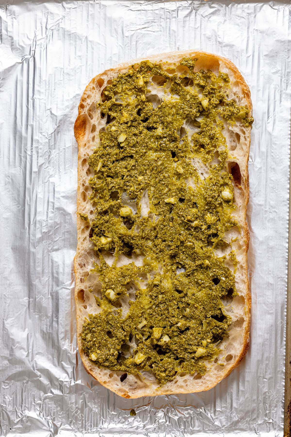 Pesto sauce on sliced rustic bread on a baking sheet with foil paper.