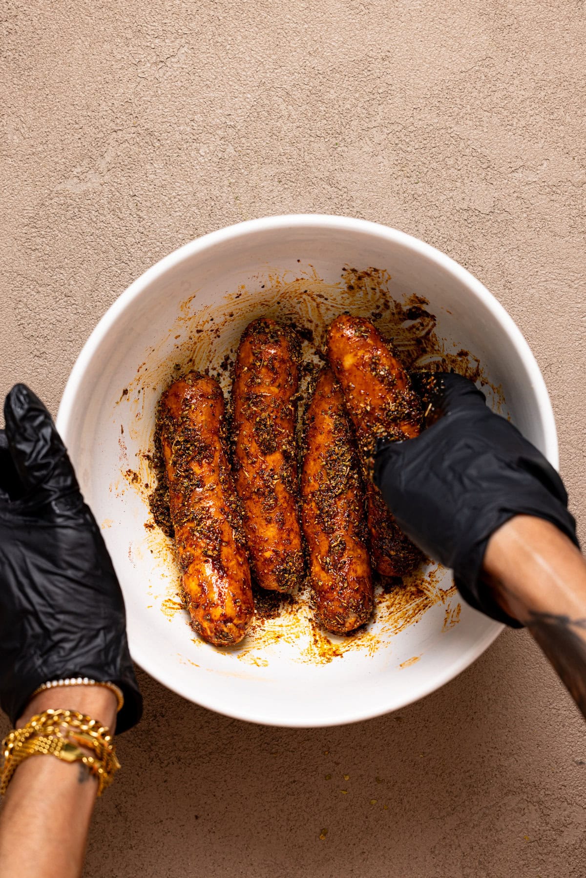 Sausages being seasoned with hands in black gloves in a bowl.