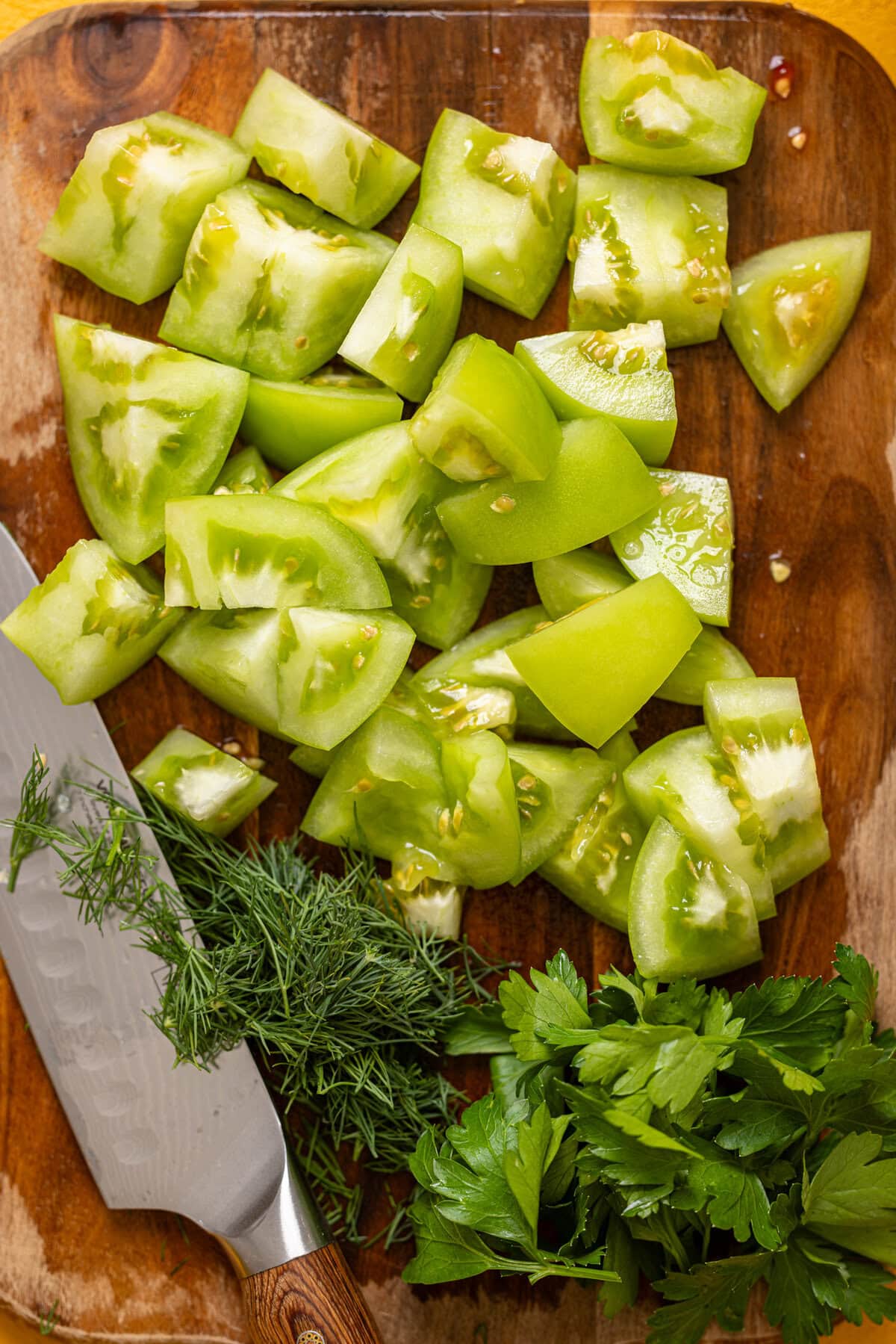 Green tomatoes and herbs chopped on a cutting board with a knife.