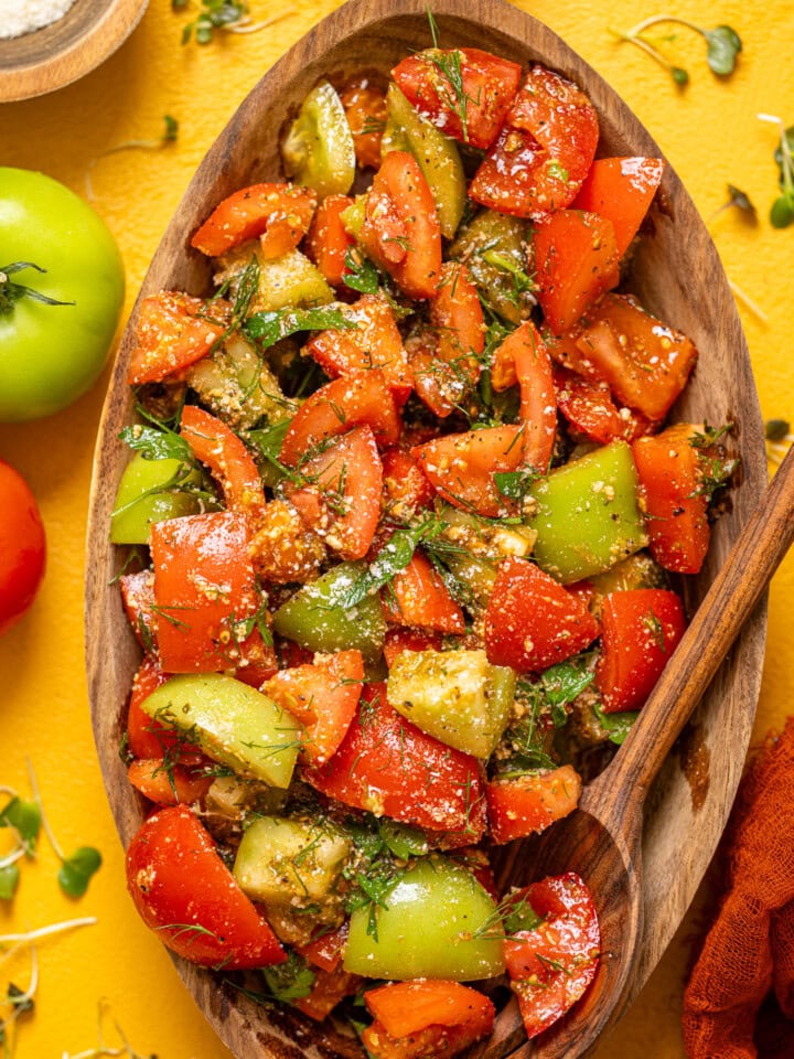 Tomato salad in a wooden serving bowl with a spoon and tomatoes on a yellow table.