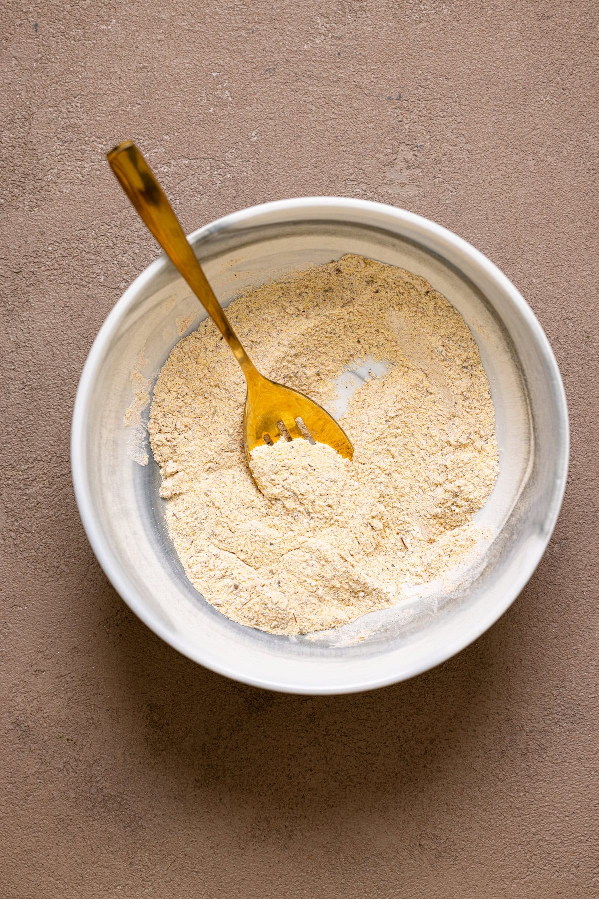 A marble bowl with seasoned flour and a spoon on a brown grey-ish table.