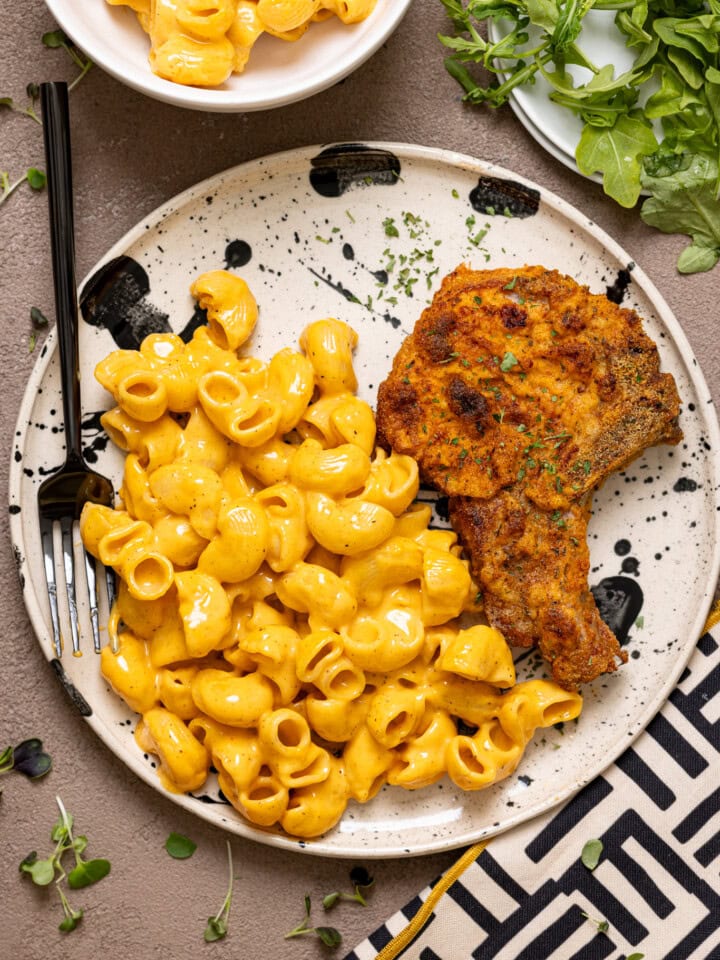 Plate with porks chops and mac and cheese with a fork and side of greens.