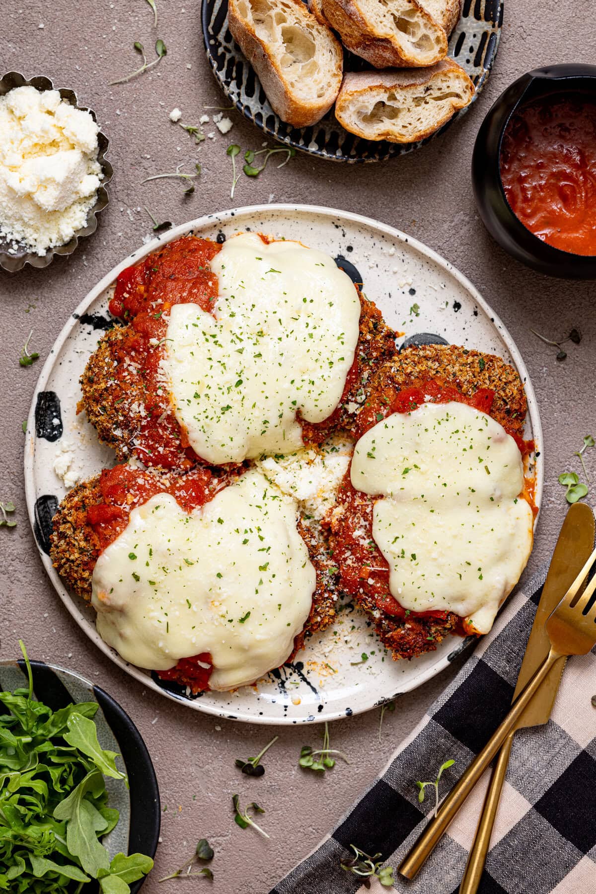 Chicken Parmesan on a plate with a fork, knife, vodka sauce, parmesan, bread, and a side of greens.