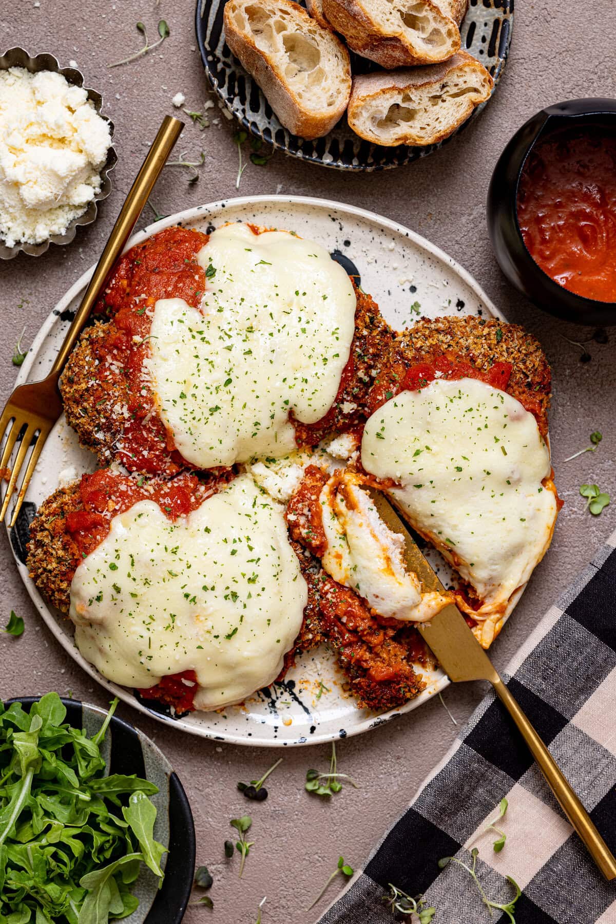 Chicken parmesan on a plate with a fork and knife and a side of parmesan and bread.