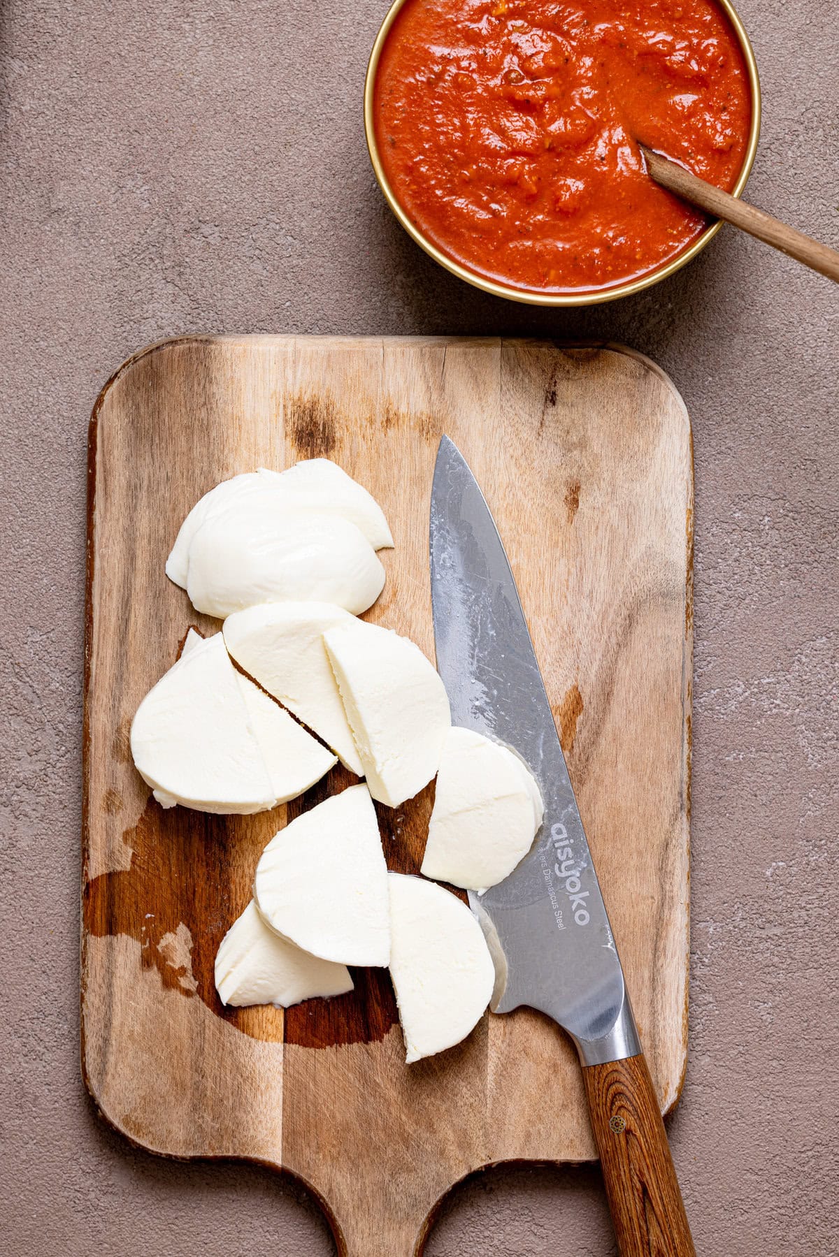 Sliced mozzarella cheese on a cutting board with a knife and a bowl with vodka sauce.