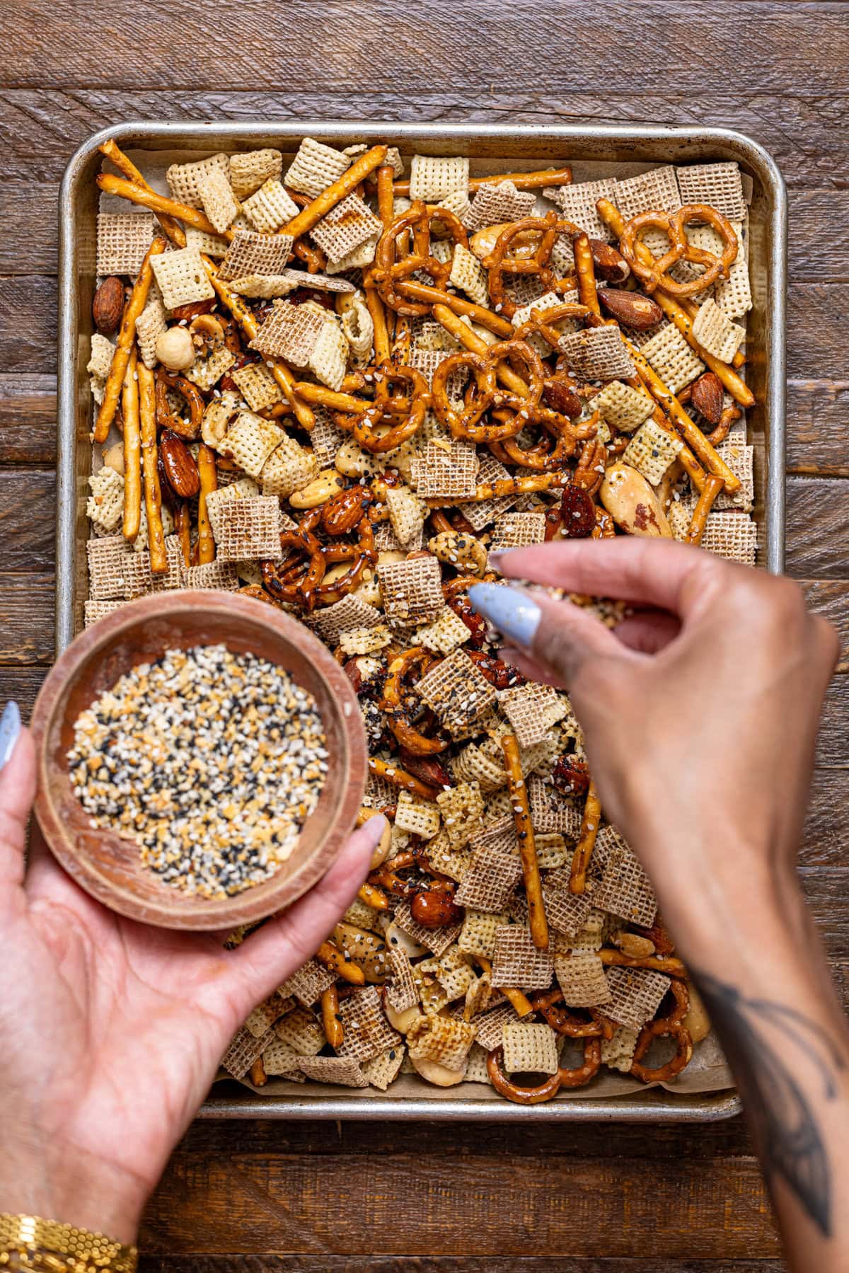 Chex mix on a baking sheet with everything bagel seasoning being sprinkled with hands.