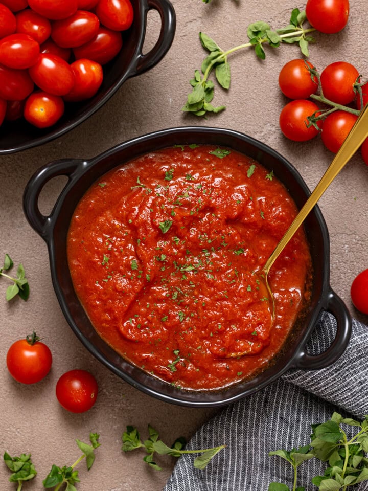 Tomato sauce in a black bowl with a spoon, tomatoes, and herbs on a brown grey-ish table.