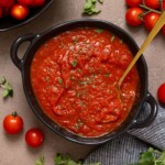 Tomato sauce in a black bowl with a spoon, tomatoes, and herbs on a brown grey-ish table.