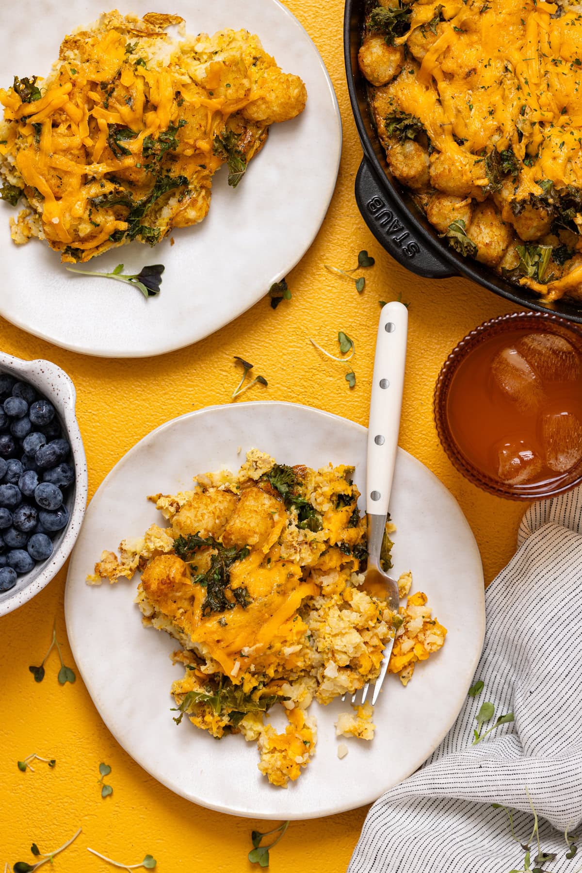 Two plates of tater tot casserole with a fork and a drink on a yellow table.