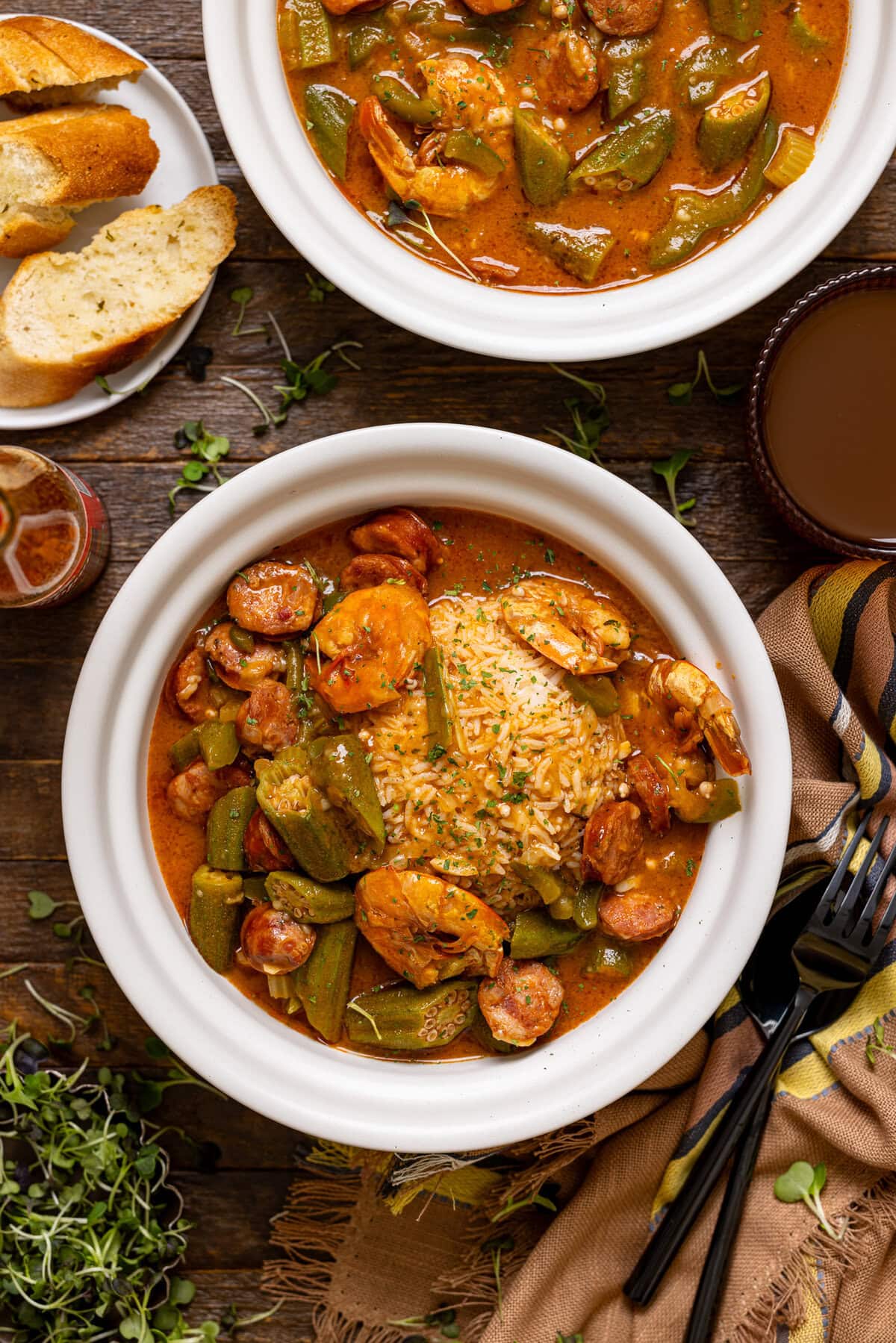 Two bowls of gumbo with a drink, side of bread, and fork and spoon.