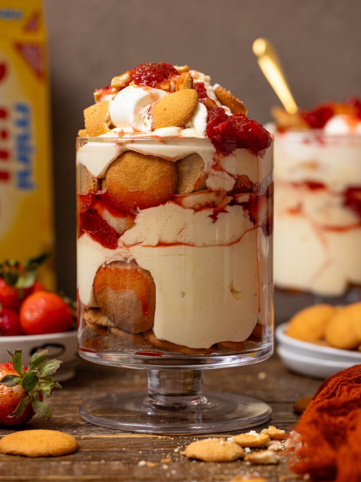 Two trifle bowls of strawberry banana puddings with a box of nilla wafers and strawberries in the background.