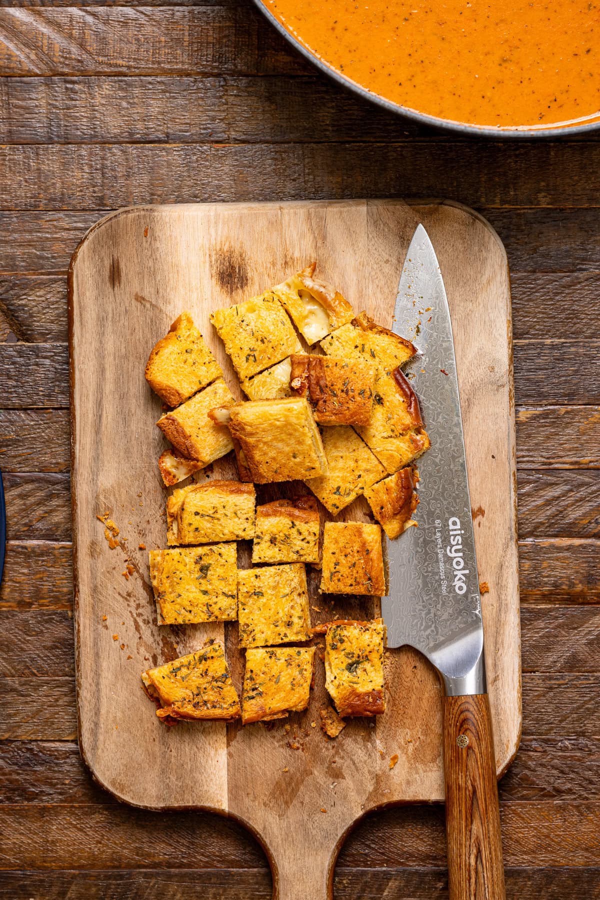 Grilled cheese cut into crouton squares on a cutting board with a knife.