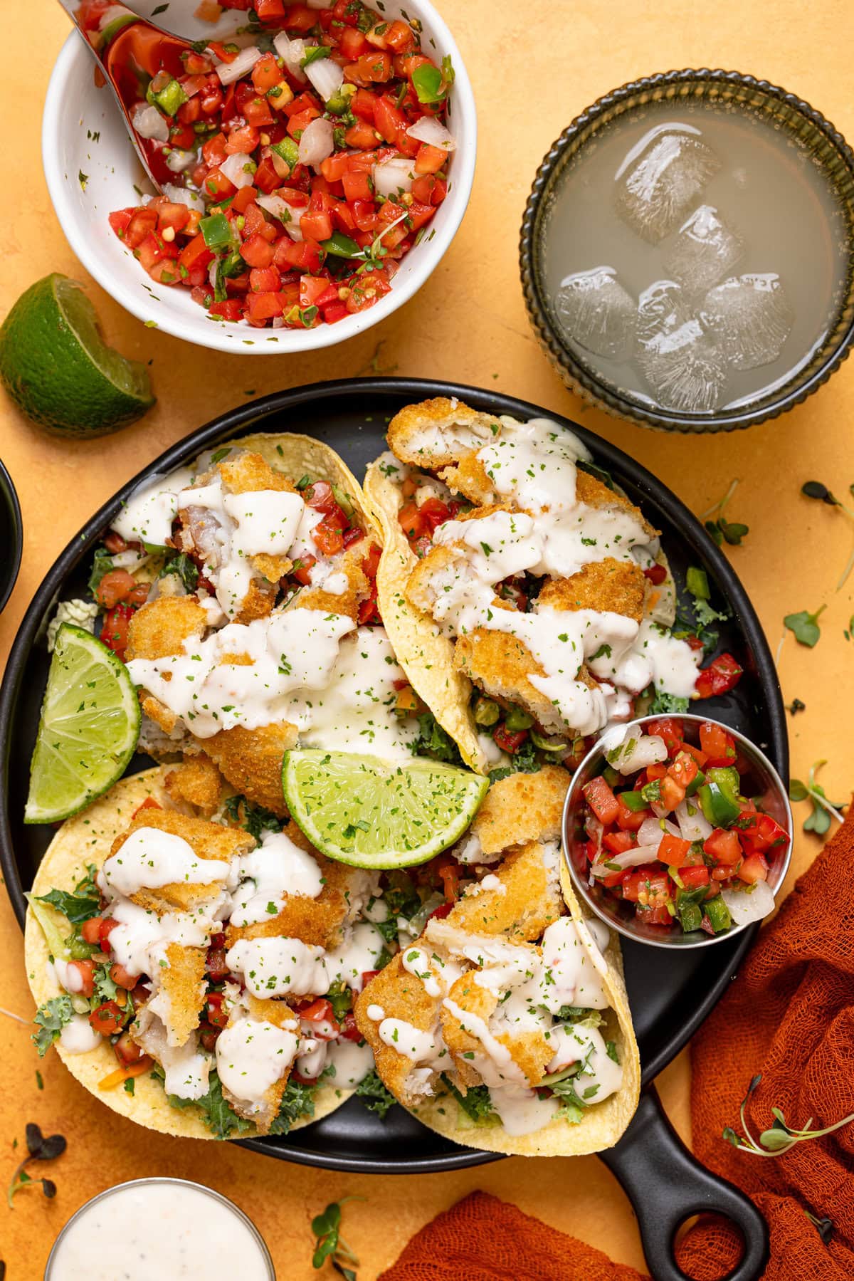 Fish Baja tacos on a platter with toppings and a drink on a yellow table.