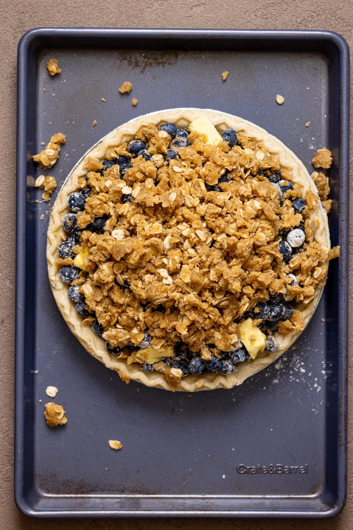 Crumble topping atop blueberry pie ingredients on a blue baking sheet.