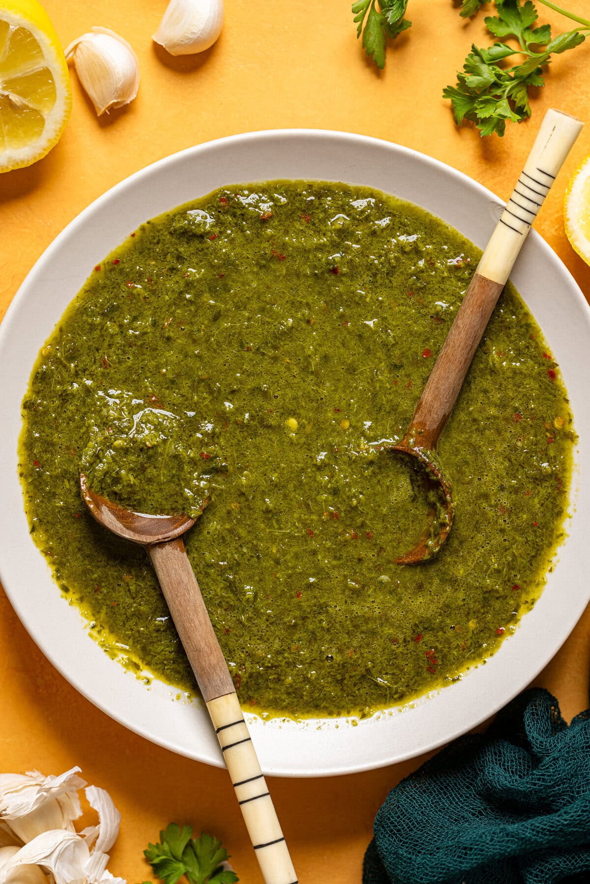 Chimichurri sauce in a white bowl on a yellow table with ingredients. 