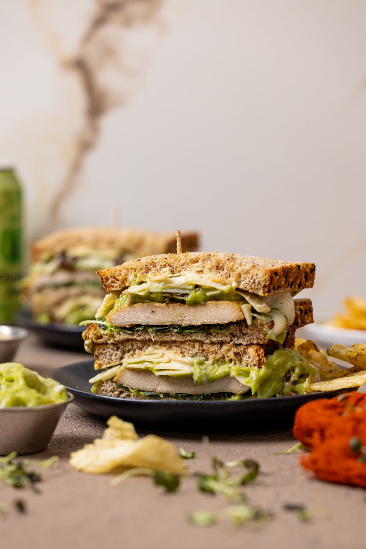 Sandwiches stacked on black plates with chips, mashed avocado, and a drink.