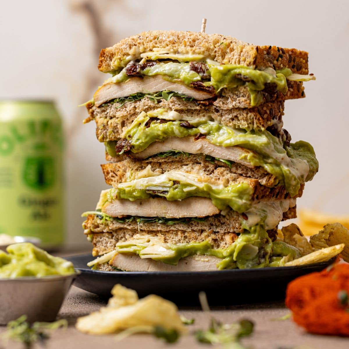 Chicken avocado sandwiches stacked on a plate with a drink and chips.