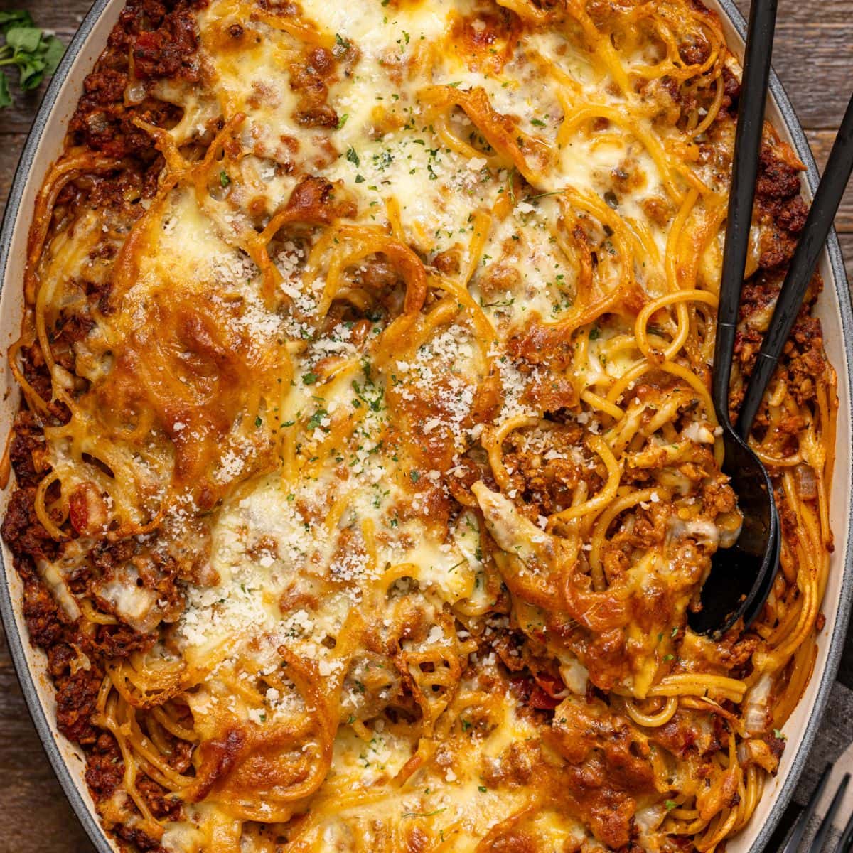 Baked spaghetti in a baking dish with two serving spoons on a brown wood table.