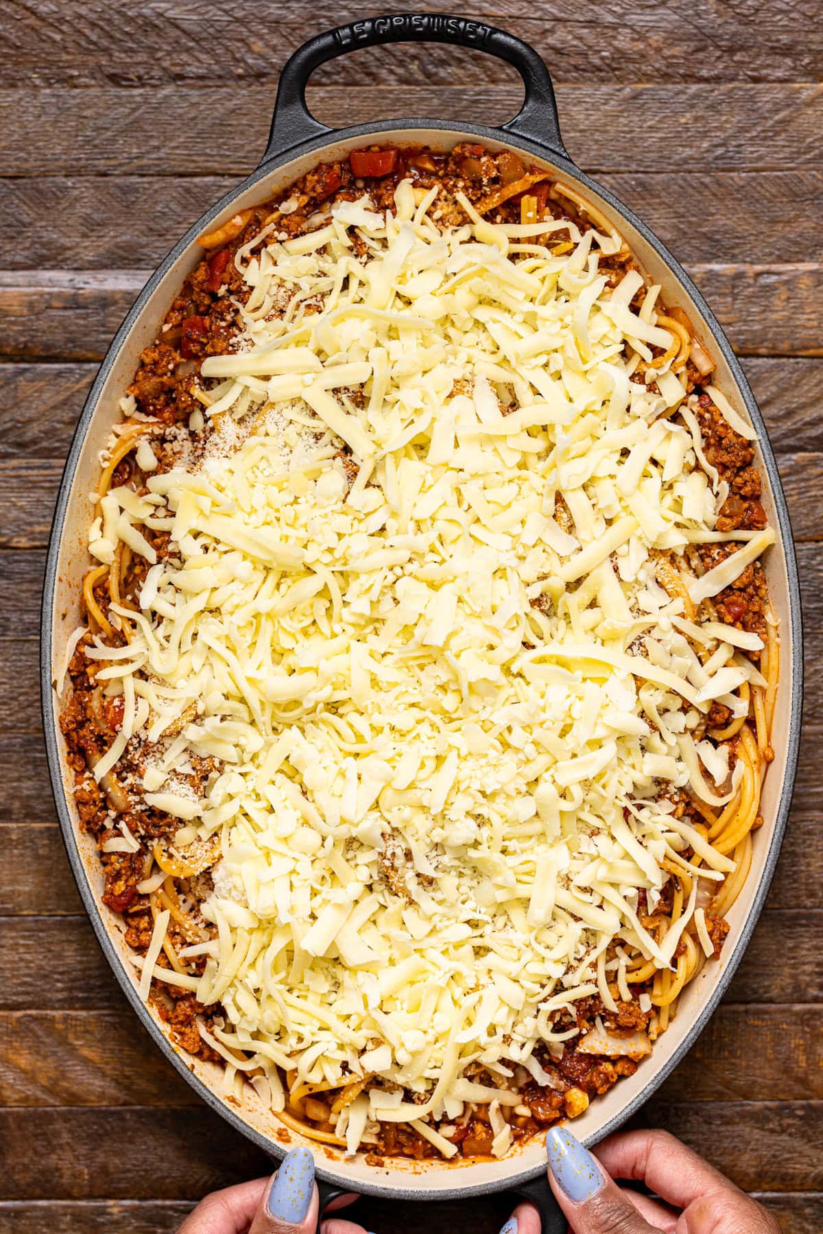 Cheese layered atop ingredients in a baking dish being held by hands.