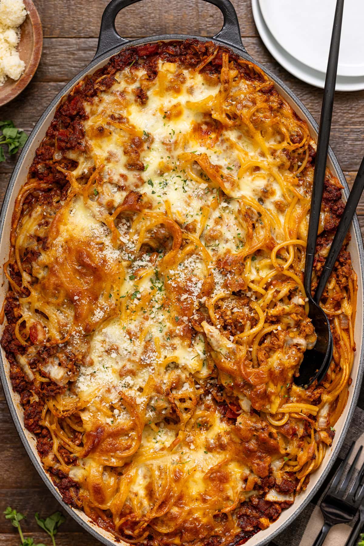 Baked spaghetti in a baking dish with two serving spoons on a brown wood table.