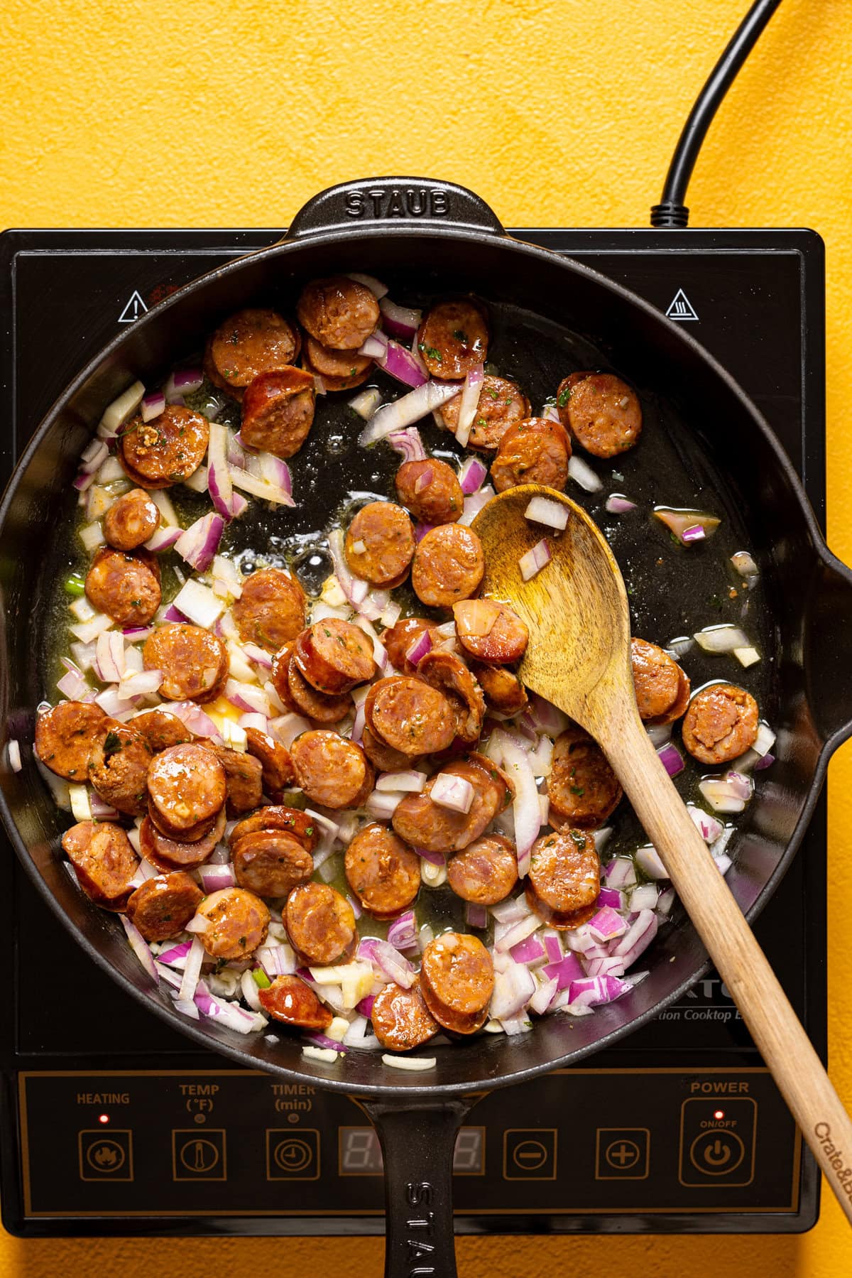 Sliced sausage and onions being sautéed in a skillet with a spoon.