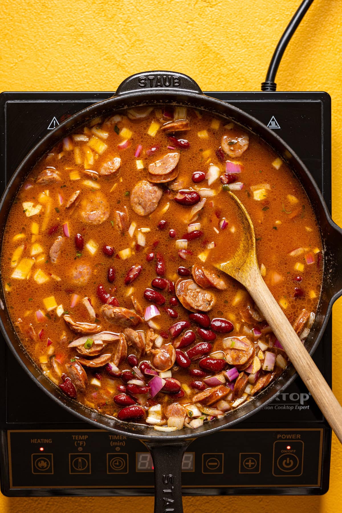 Red beans and sausage being cooked in a skillet over heat being stirred with a wooden spoon.