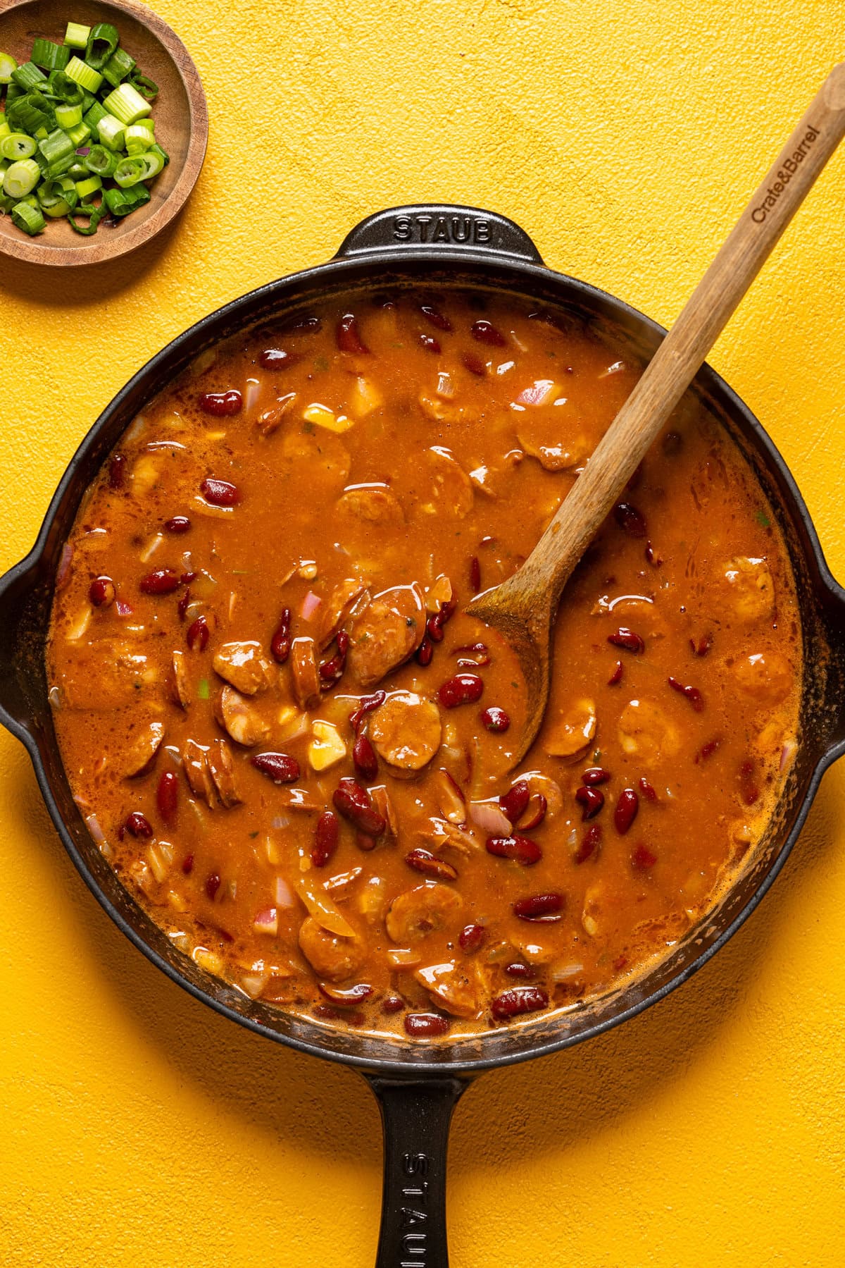 Cooked red beans in a skillet on a yellow table with a wooden spoon.