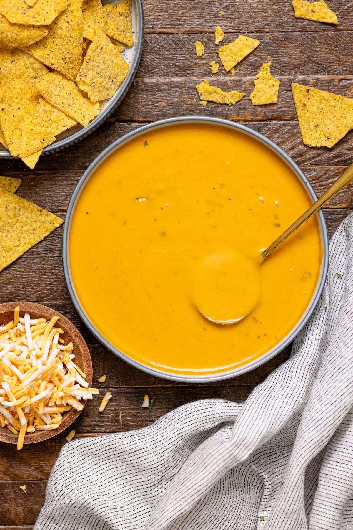 Bowl of nacho cheese with a spoon, shredded cheese, and tortilla chips.