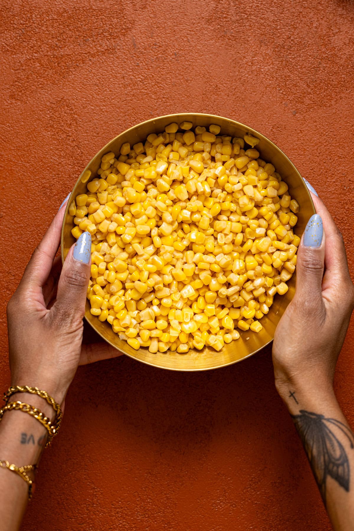 A bowl of corn kernels being held with hands on a burnt orange table.