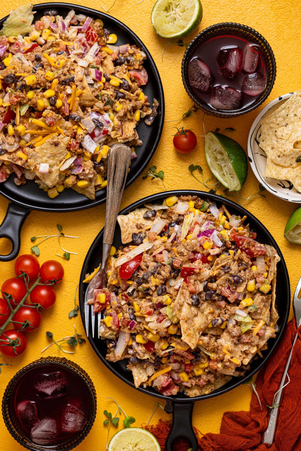 Two plates of 7-layer taco salad with a fork, drinks, tomatoes, and limes on the side.