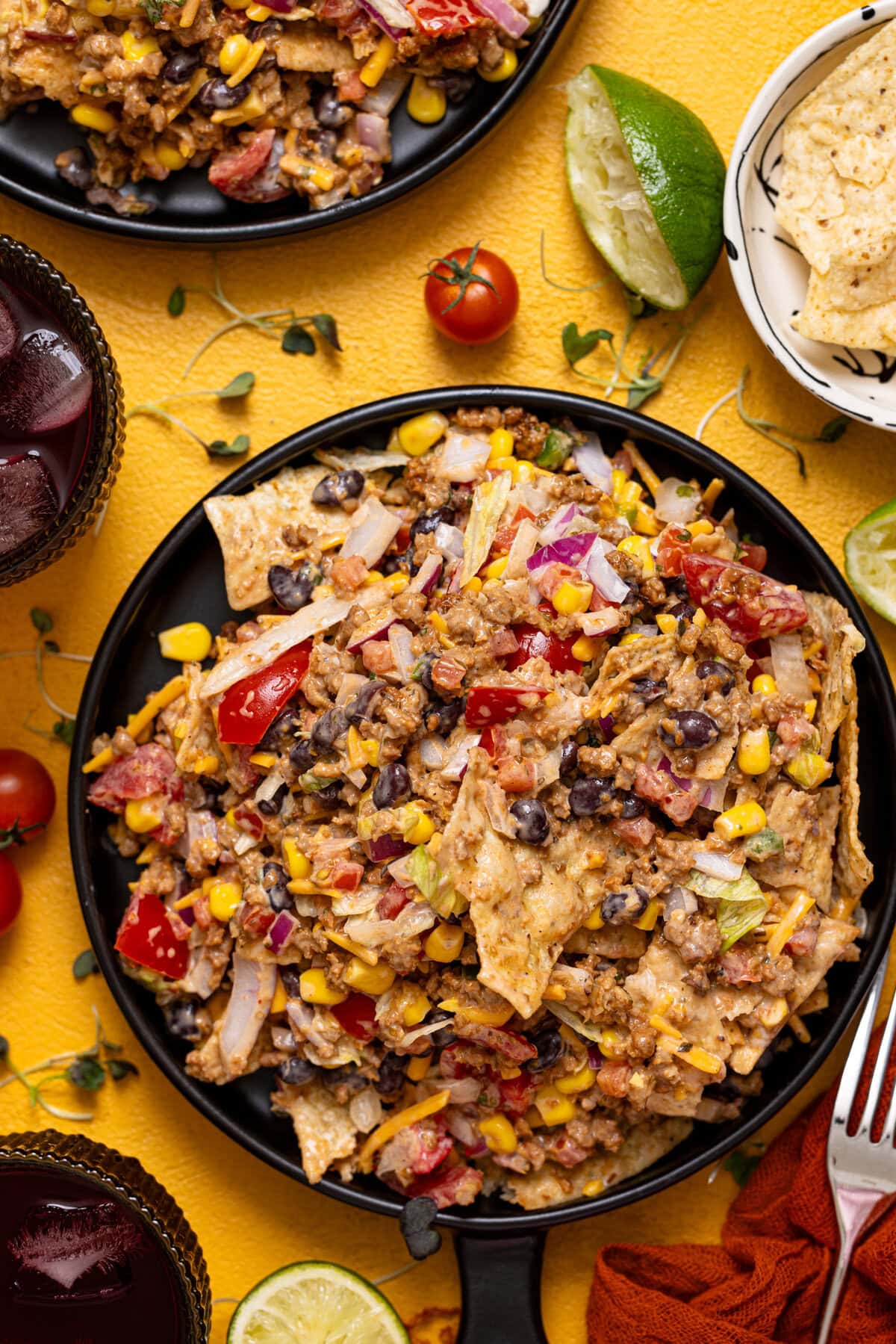 Up close shot of 7-layer taco salad on a black plate with drinks, lime, and a fork.