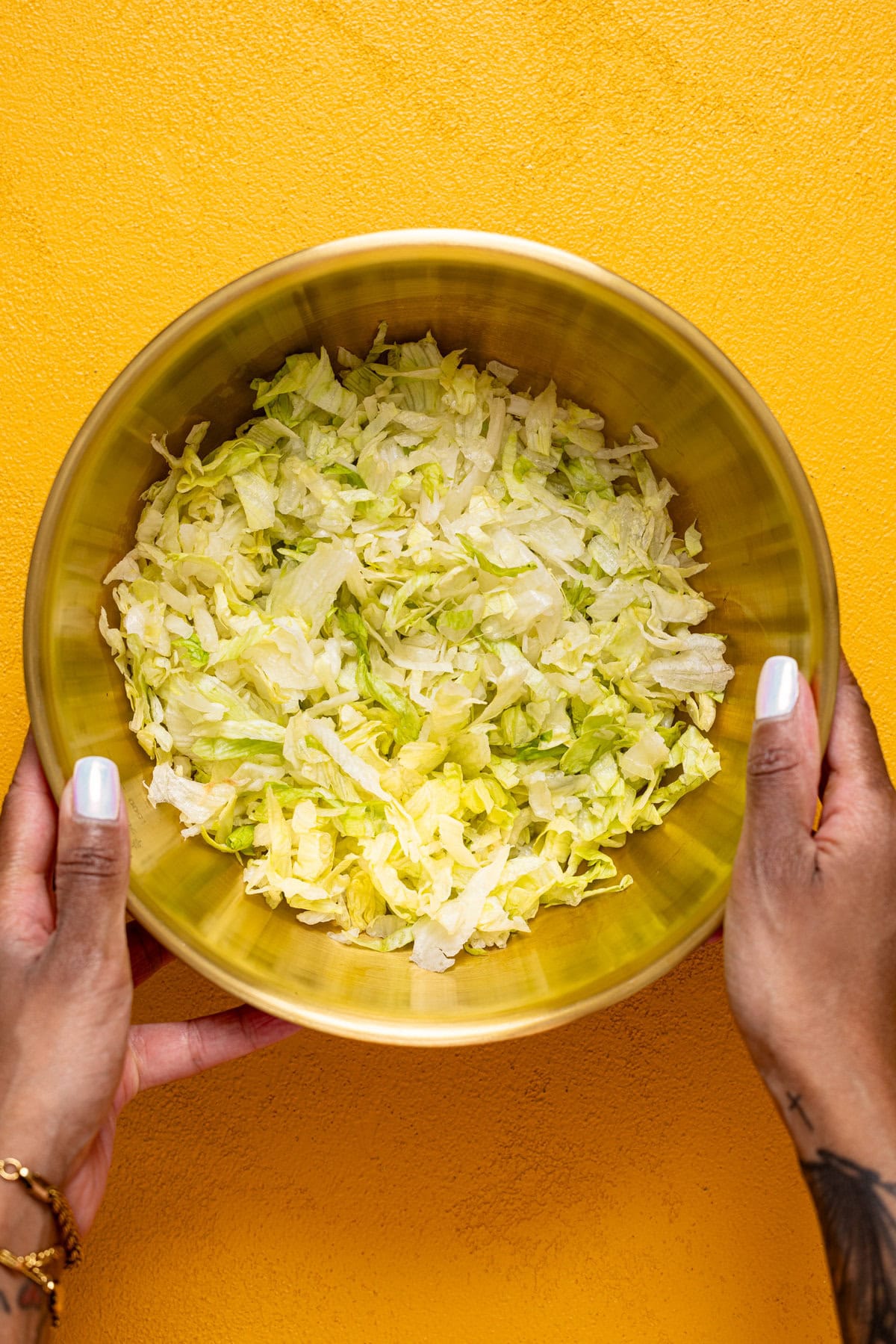 Chopped lettuce in a gold bowl being held on a yellow table.