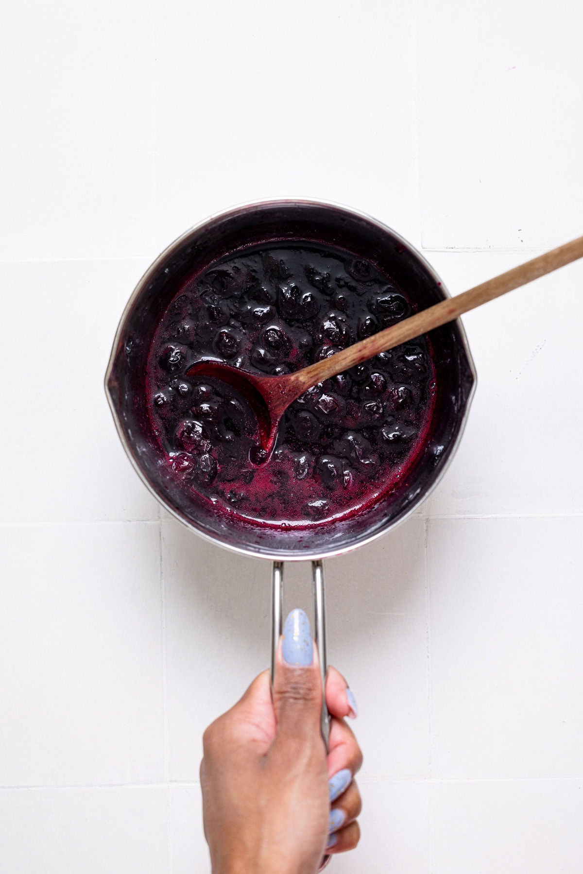 Blueberry compote in a saucepan being held on a white tile table with a wooden spoon.