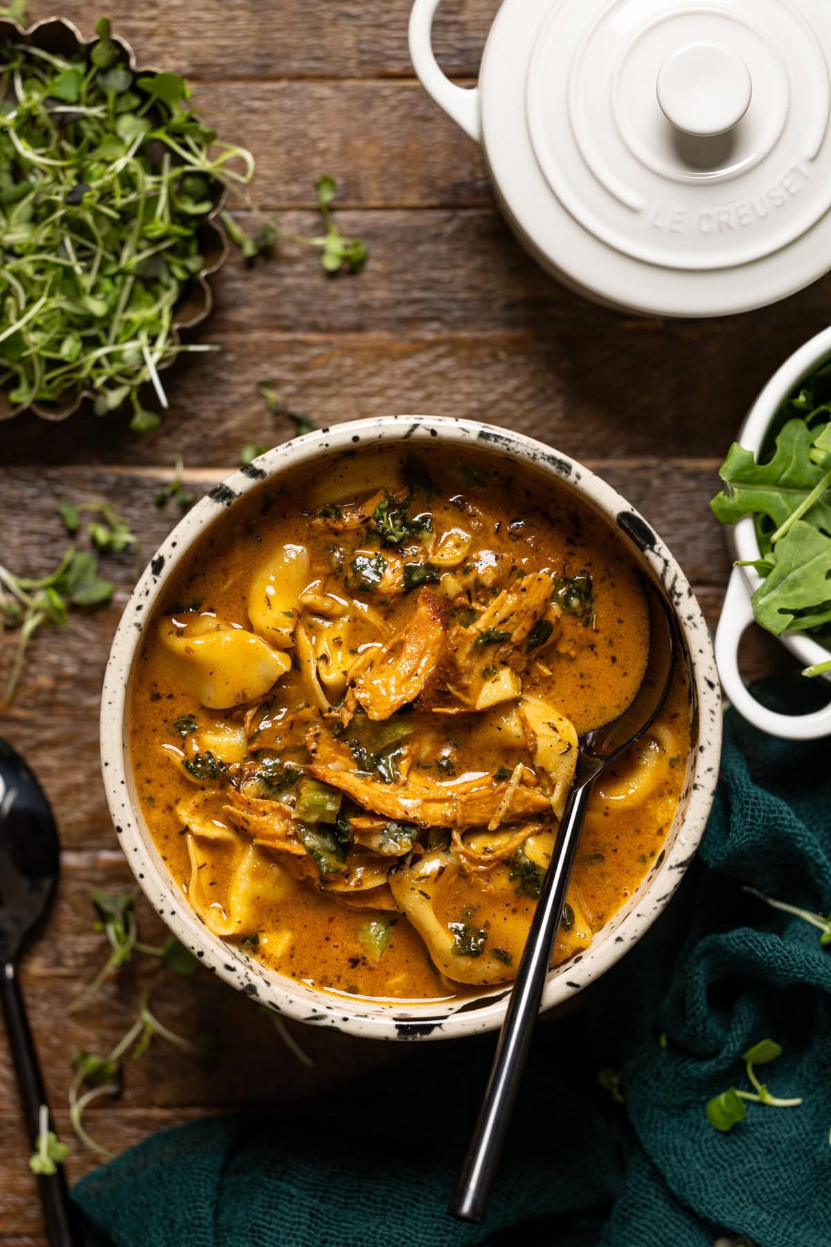 A bowl of chicken tortellini soup with greens and spoons.