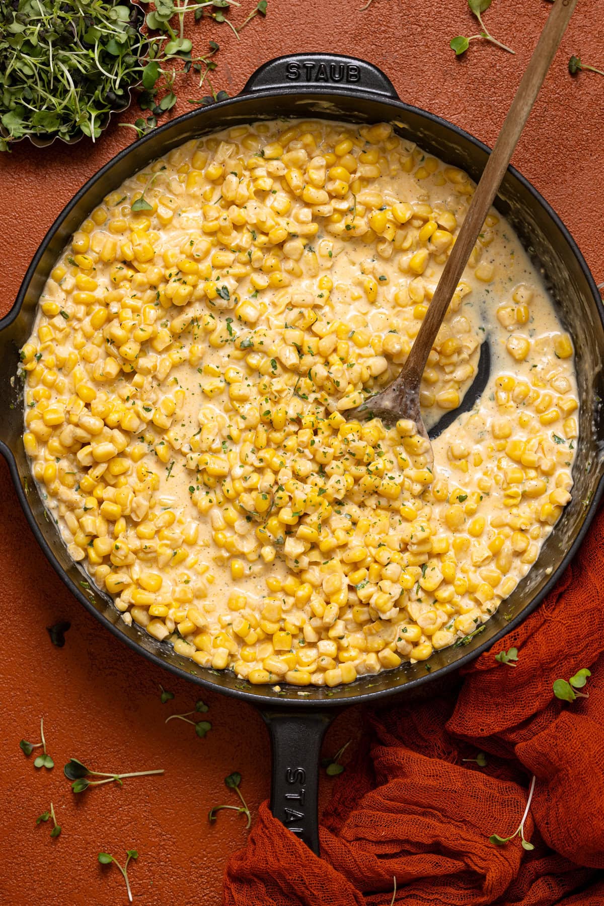 A skillet with creamed corn and a wooden spoon on a burnt orange table.