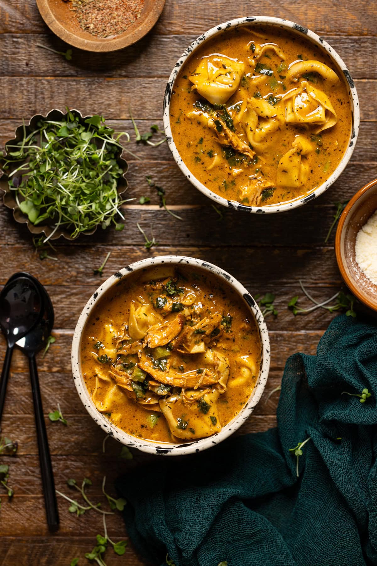 Two bowls of chicken tortellini soup on a brown wood table with spoons and greens.