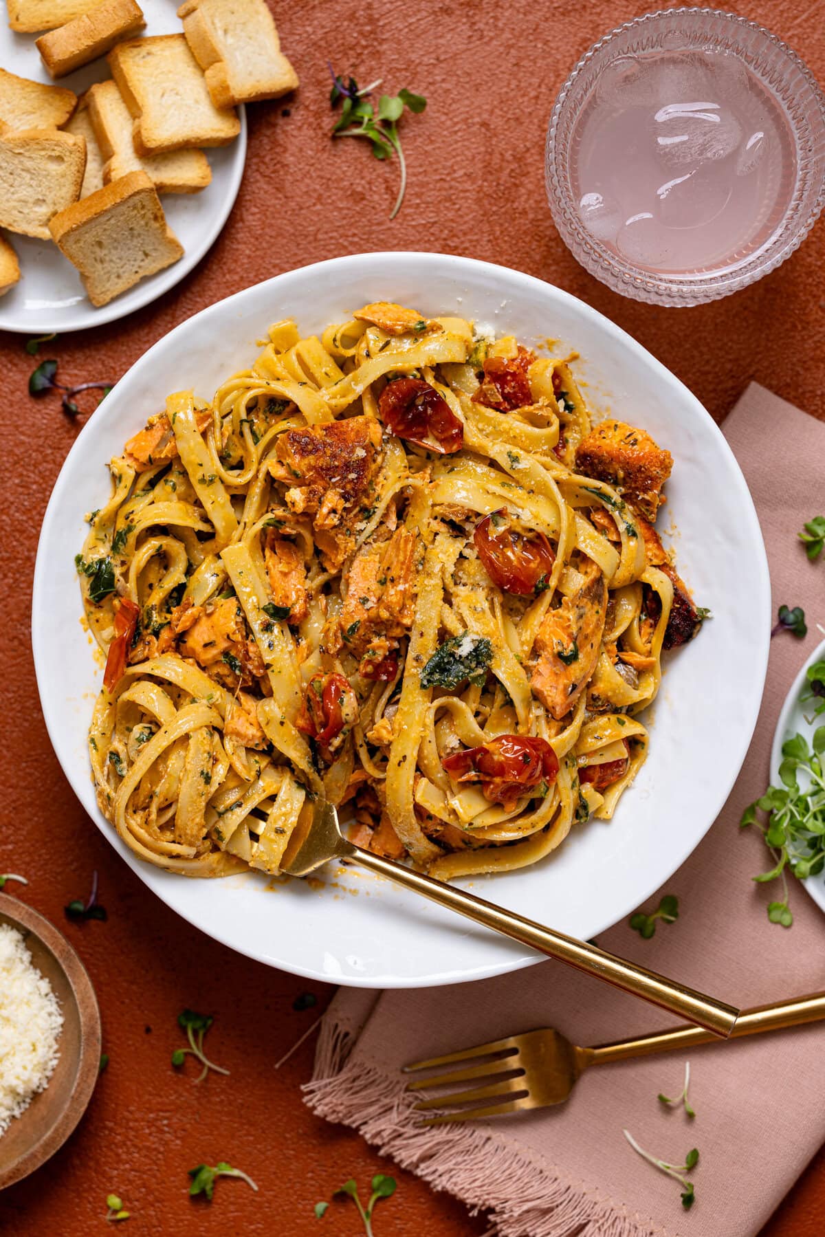 A plate of salmon pasta with forks, a drink, and side of bread.