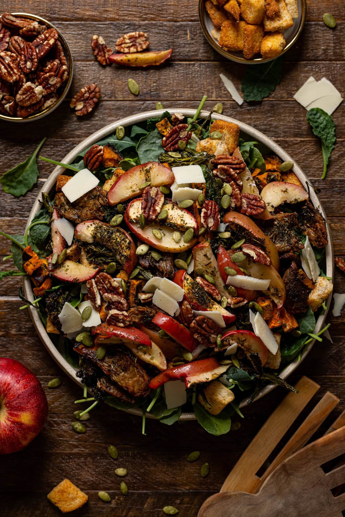 A large salad in a bowl with serving utensils, apples, pecans, and croutons. 